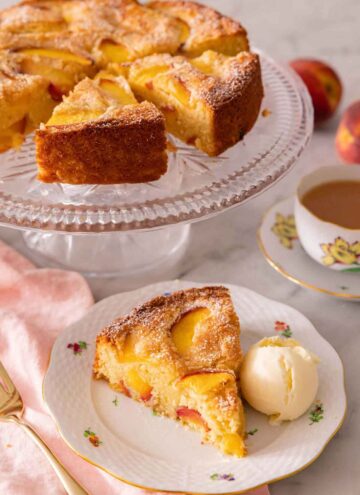 A slice of peach cake with a scoop of ice cream by a cake stand with the rest of the peach cake.