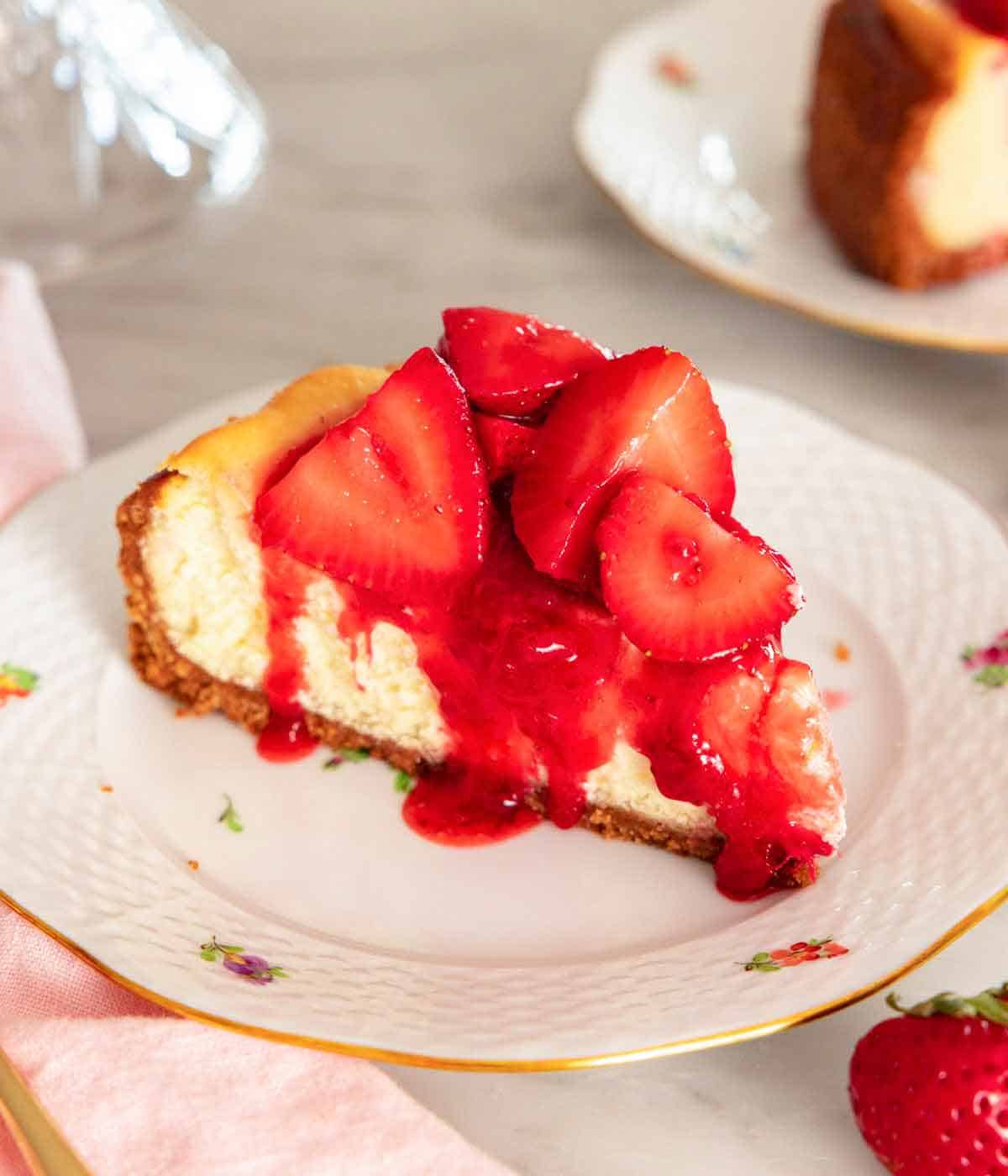 A slice of strawberry cheesecake with syrup under cut strawberries drizzling down the side of the slice, on a plate.