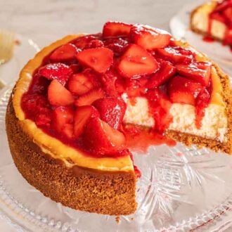 Overhead view of a strawberry cheesecake on a clear cake stand with a piece cut out.