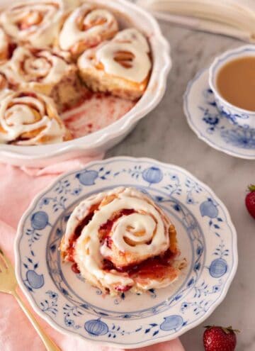 A plate with a serving of strawberry roll with cream cheese icing on top, by a baking dish with the rest of the rolls.