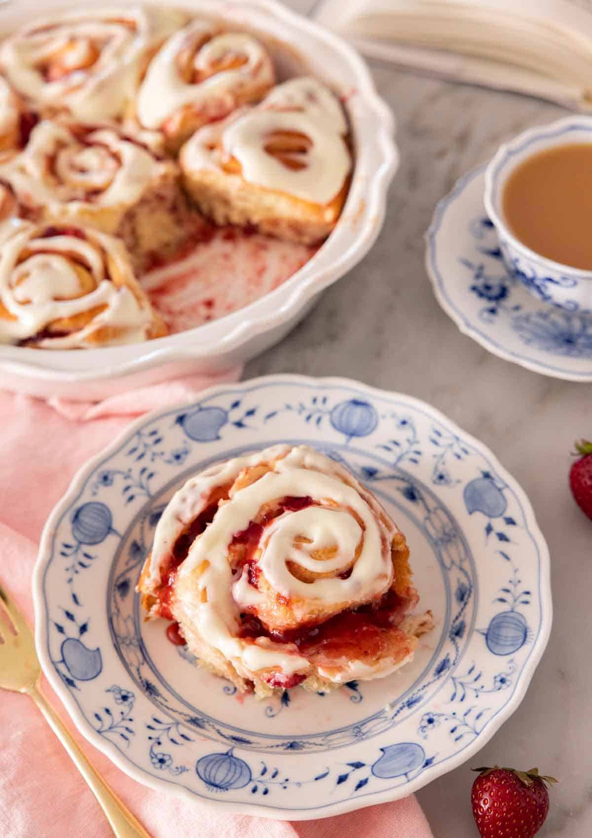 A plate with a serving of strawberry roll with cream cheese icing on top, by a baking dish with the rest of the rolls.