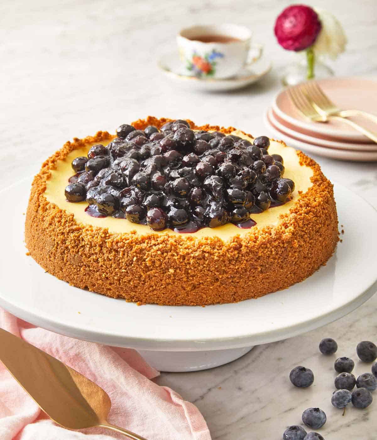 A cake stand with a blueberry cheesecake with a graham cracker crust going up the sides with blueberry sauce in the middle.