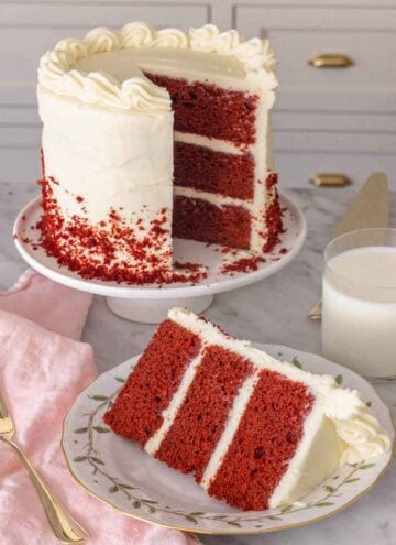 A cake stand with a slice of red velvet cake cut out and placed on a plate in front.