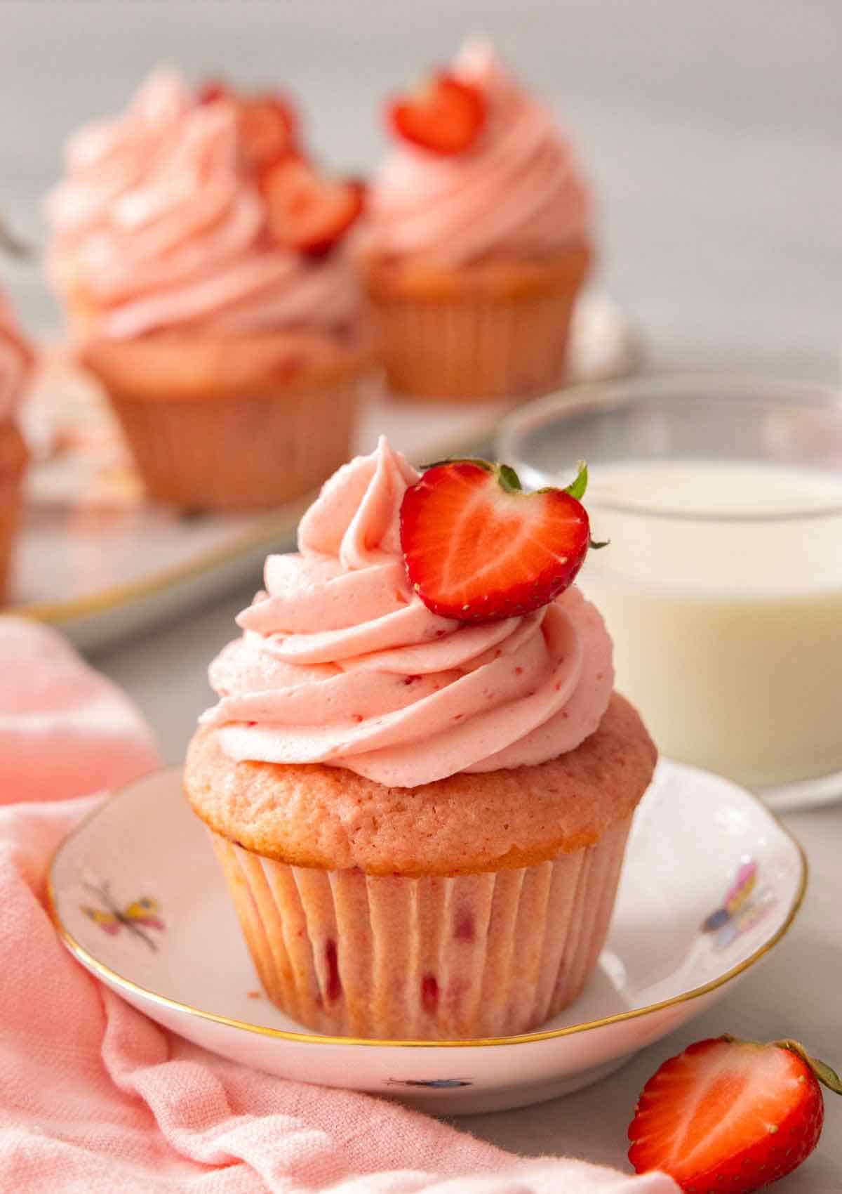 A strawberry cupcake with half a strawberry on top of the pink frosting. More cupcakes and milk in the background.