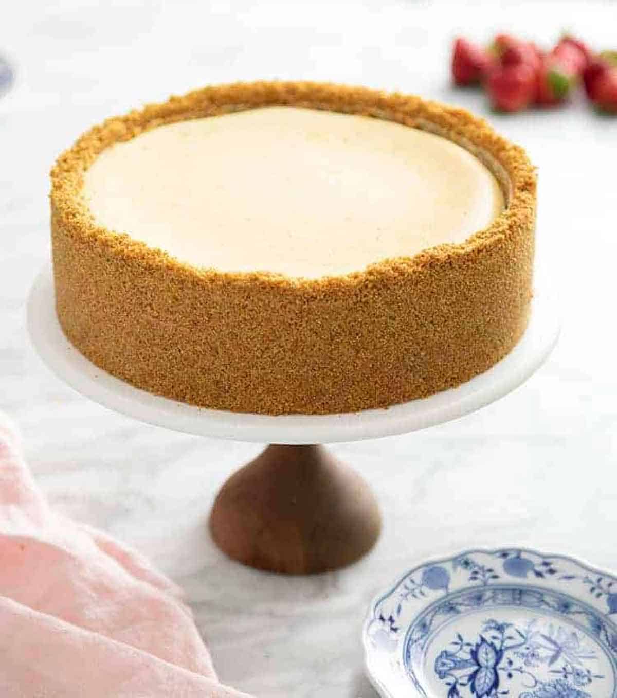A cake stand with a whole cheesecake on it with graham cracker crust going up the entire side.