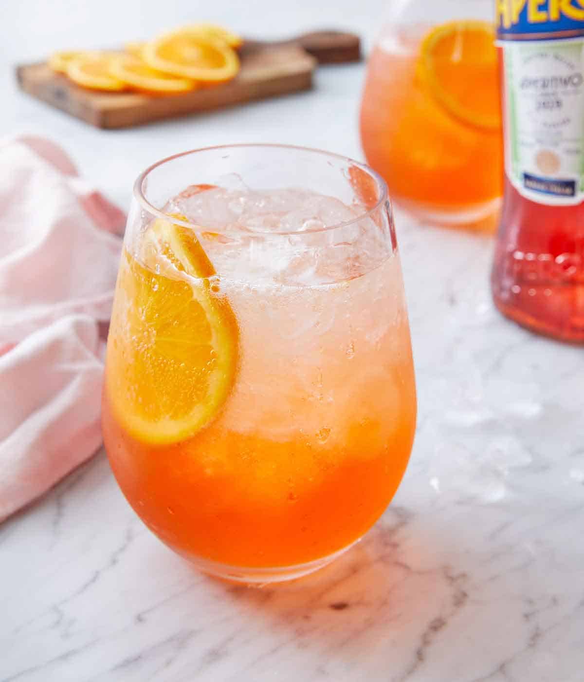 A glass of aperol spritz with a sliced orange inside and more slices in the background with another drink.
