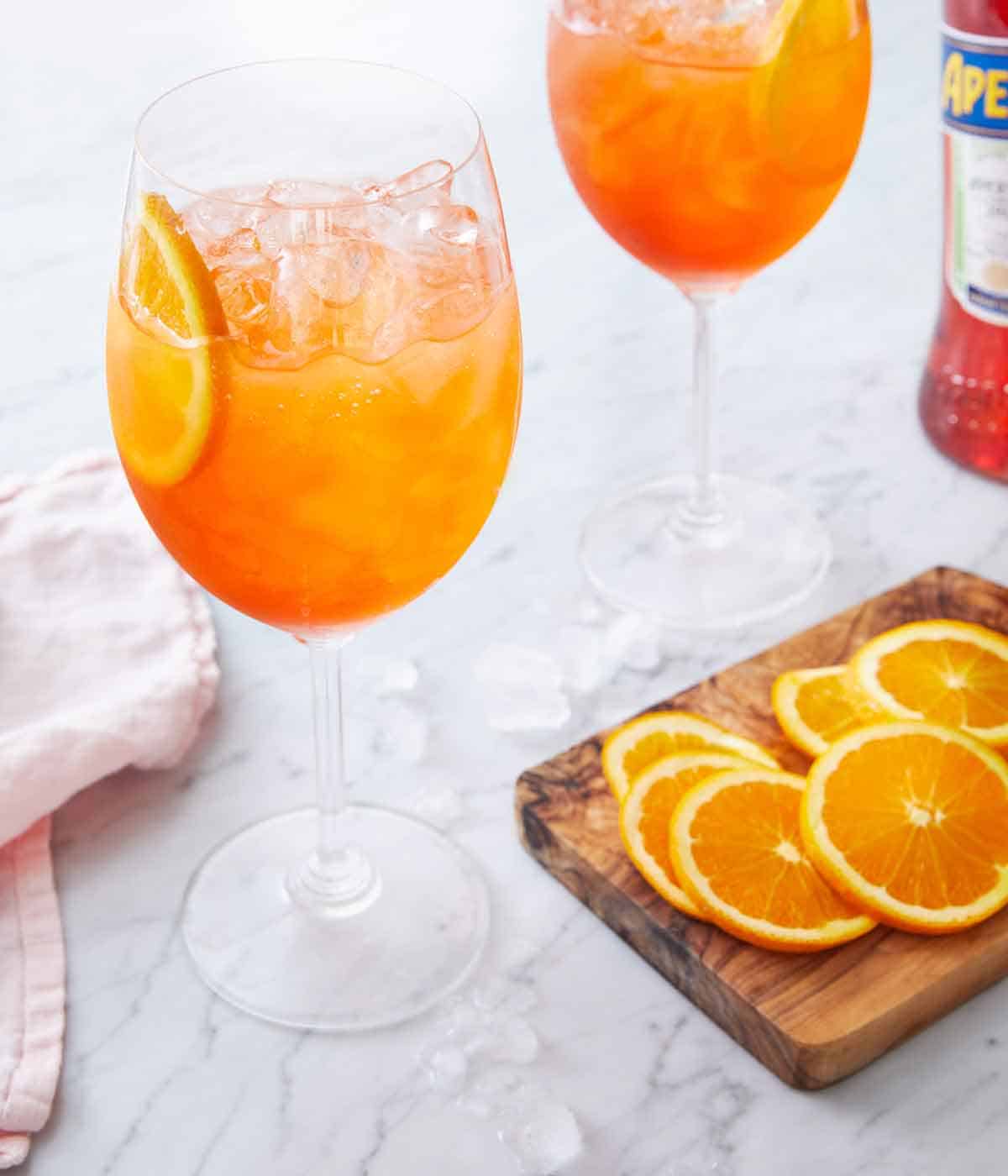 Stemmed wine glasses with aperol spritz by a cutting board with sliced oranges.