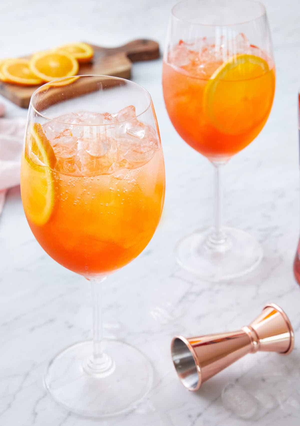 Two stemmed wine glasses of aperol spritzes with a jigger in front and sliced oranges in the background.