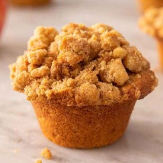 A apple muffin on a marble counter with a second muffin and an apple out of focus in the background.