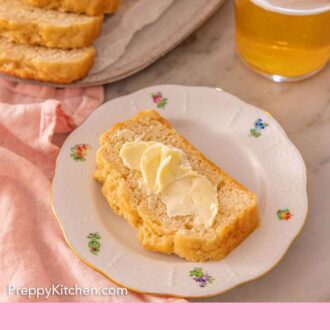Pinterest graphic of a plate with a slice of beer bread with butter slathered on top with more bread and a drink in the background.