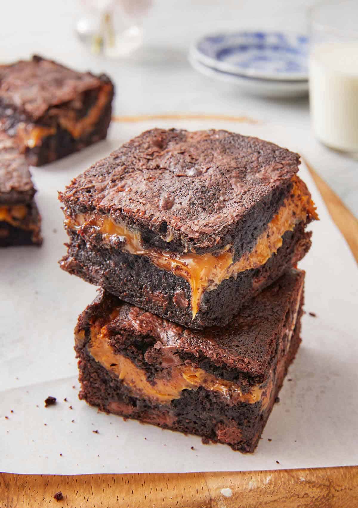 Two square pieces of caramel brownies stacked on top of each other on top of a lined cutting board.