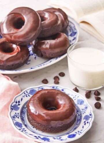 A plate with a chocolate donut by a glass of milk and additional donuts in the back.