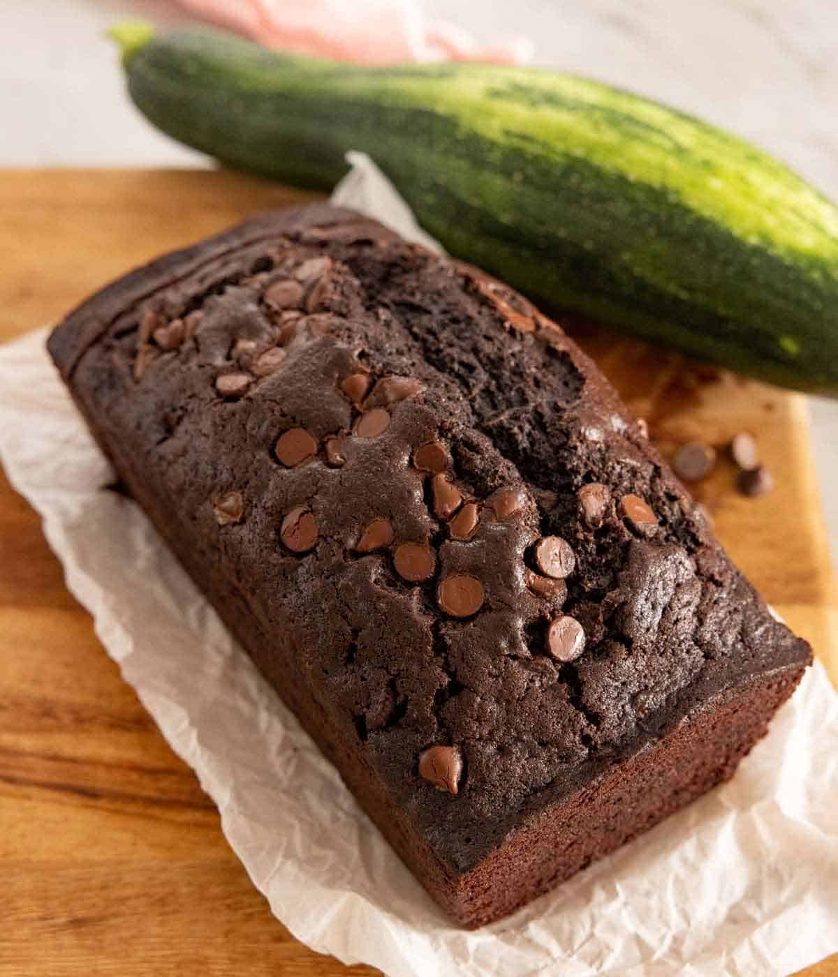 A loaf of chocolate zucchini bread with a large zucchini in the background.