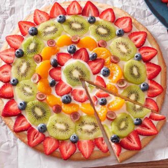 A fruit pizza placed on a parchment paper over a wooden surface, with a slice cut and slightly pulled out.