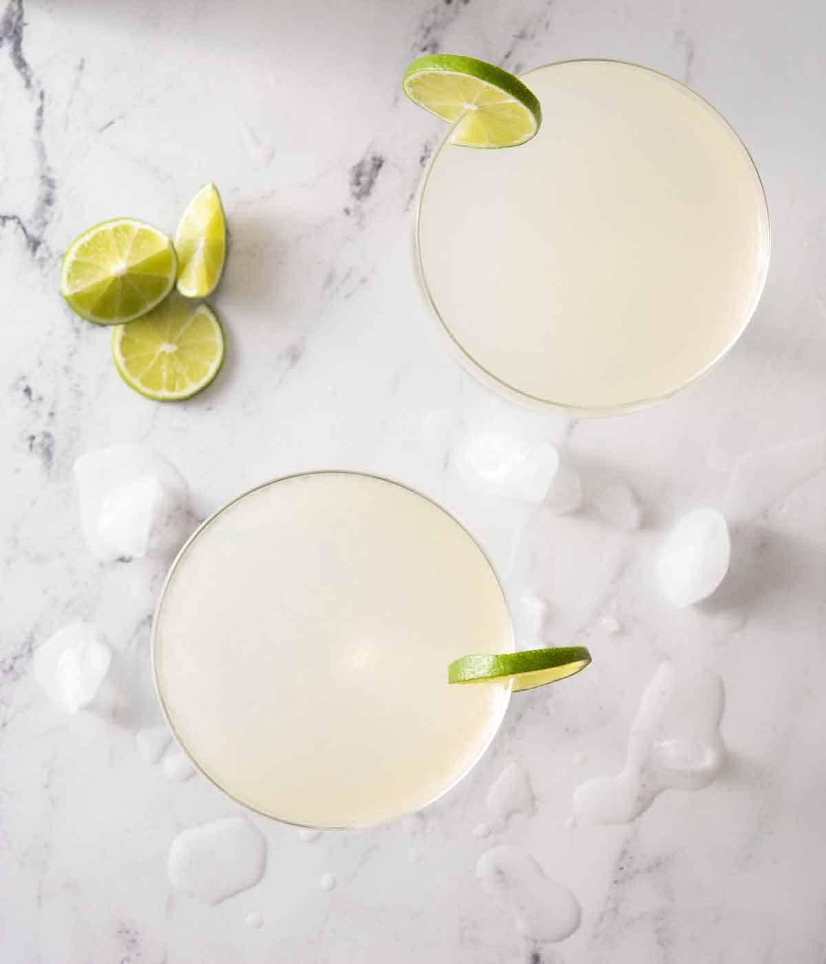 Overhead view of two glasses of gimlet cocktails with lime on the rim and on the counter along with ice.