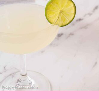 Pinterest graphic of a slightly close view of a glass of gimlet with a slice of lime on the rim and gin in the background.