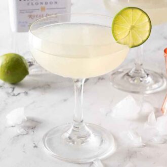 A glass of gimlet with ice, a jigger, and a whole lime behind it.