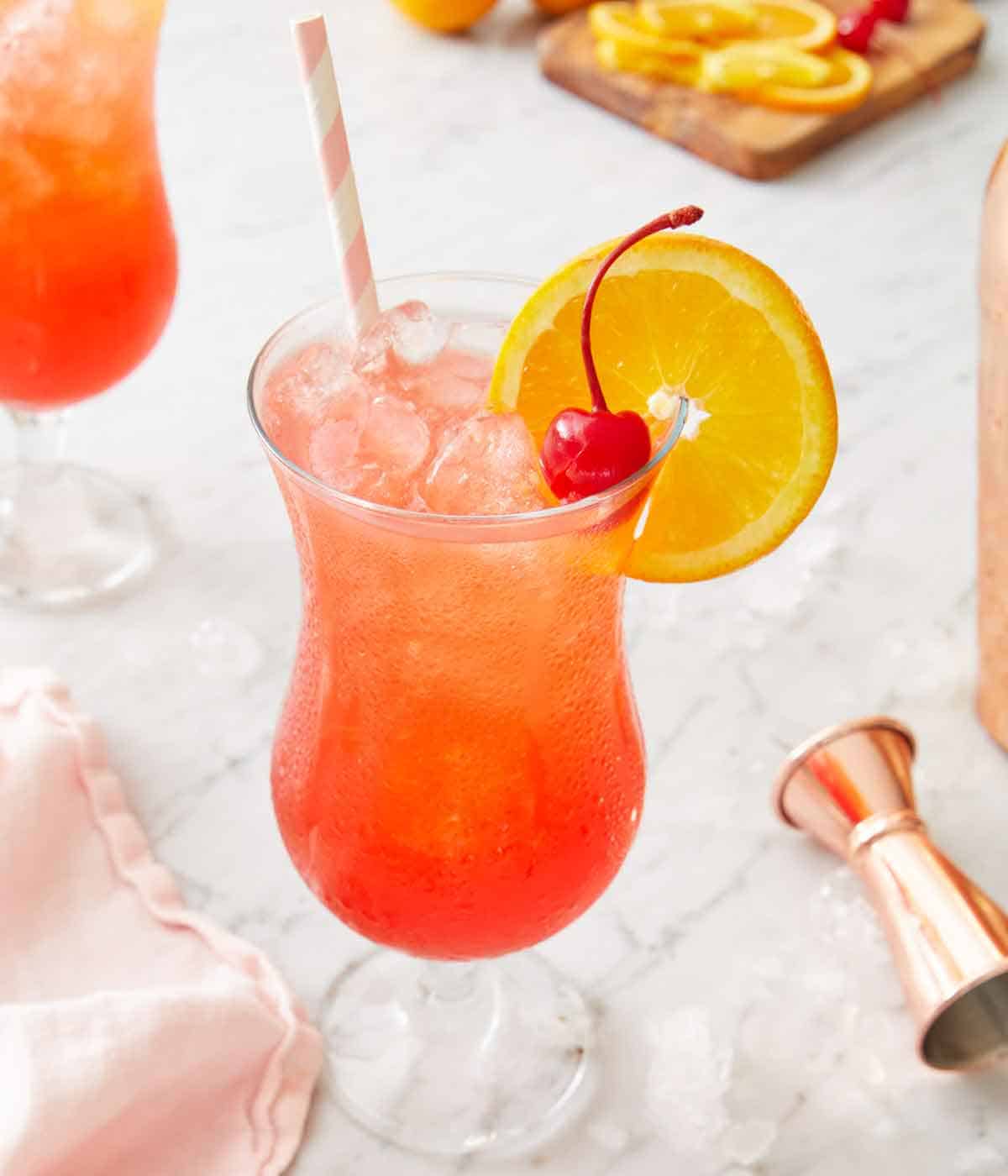A slightly overhead view of a glass of hurricane cocktail with a straw, cherry, and orange slice tucked on the rim.
