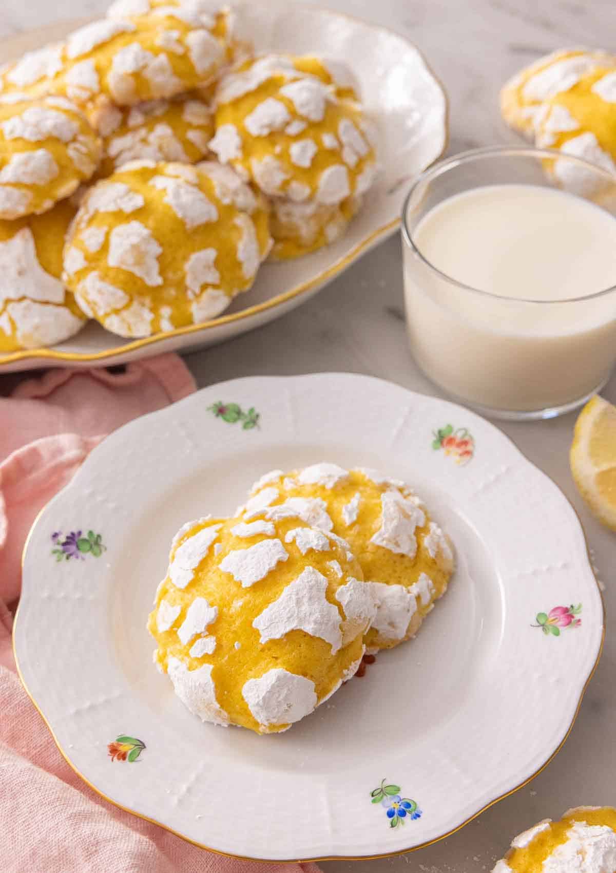 Two lemon crinkle cookies on a plate by a glass of milk in front of a platter of multiple cookies.