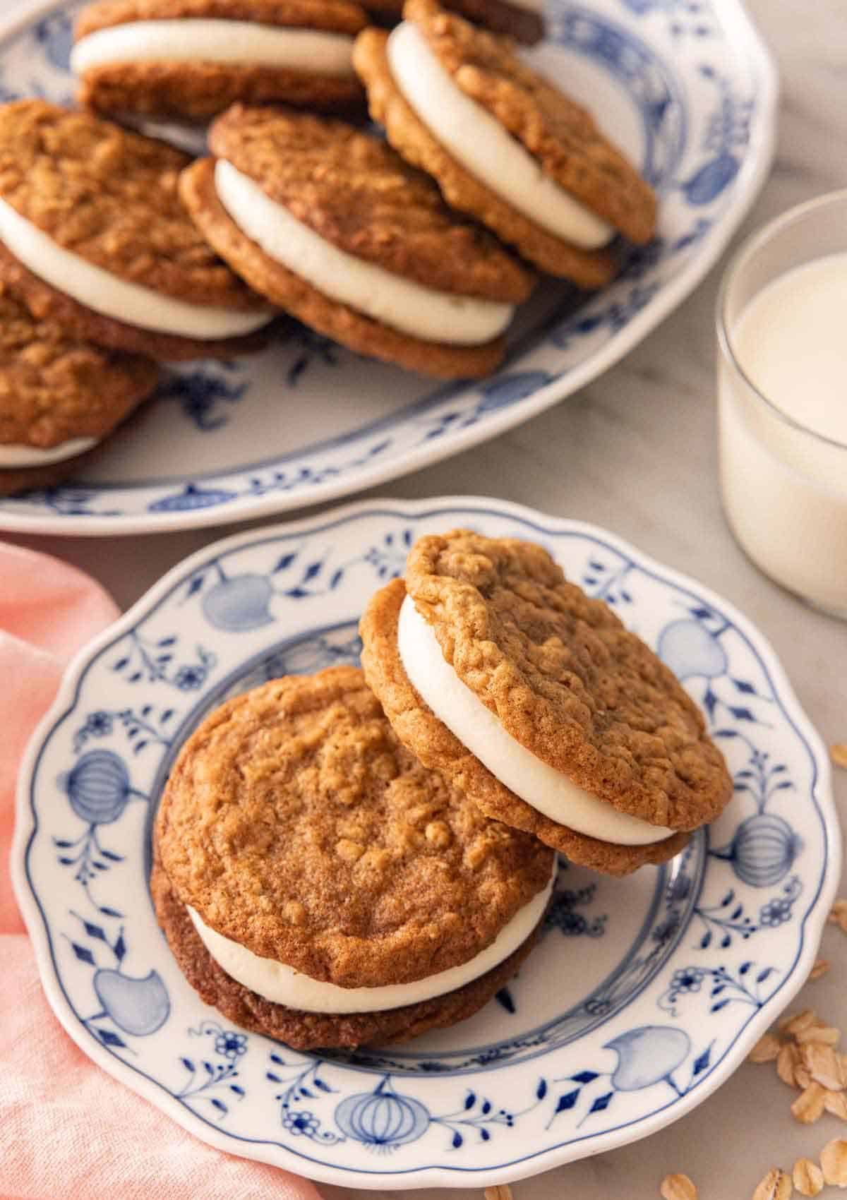 A plate with two oatmeal cream pies with a platter with more along with a glass of milk in the background.