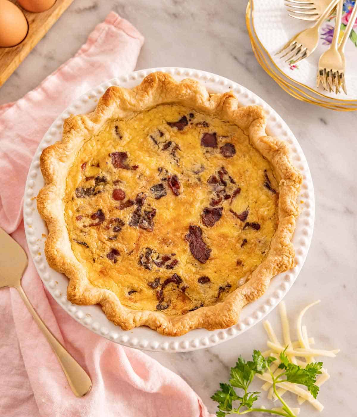 Overhead view of a quiche Lorraine with some forks, napkin, and garnish on the side.