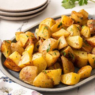 An oval platter of roasted potatoes with fresh chopped parsley on top with a stack of plates in the background.