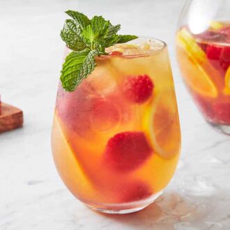 A glass of rose sangria with a pitcher in the back along with cut fruit.
