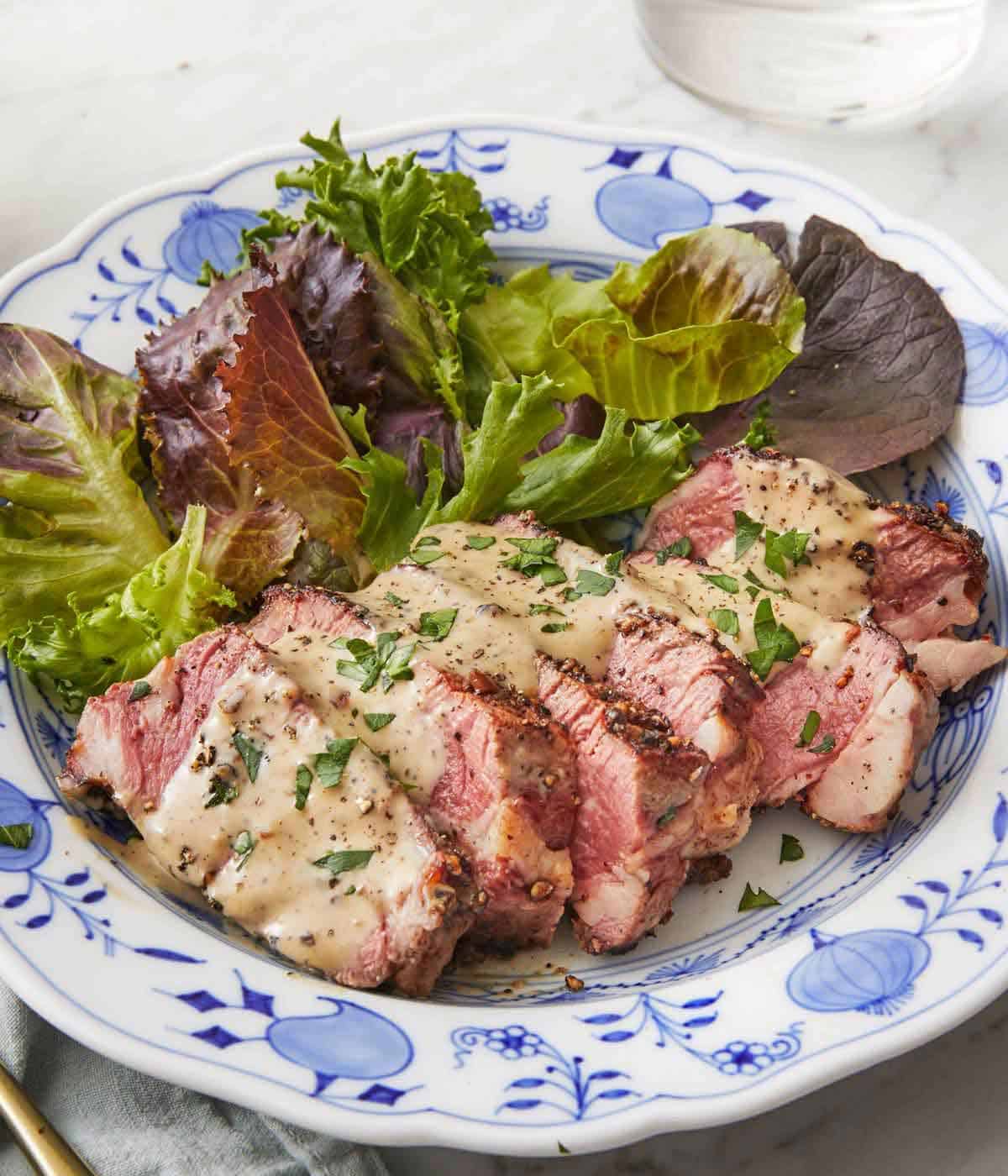 A plate of sliced steak au poivre with a side of mixed greens.