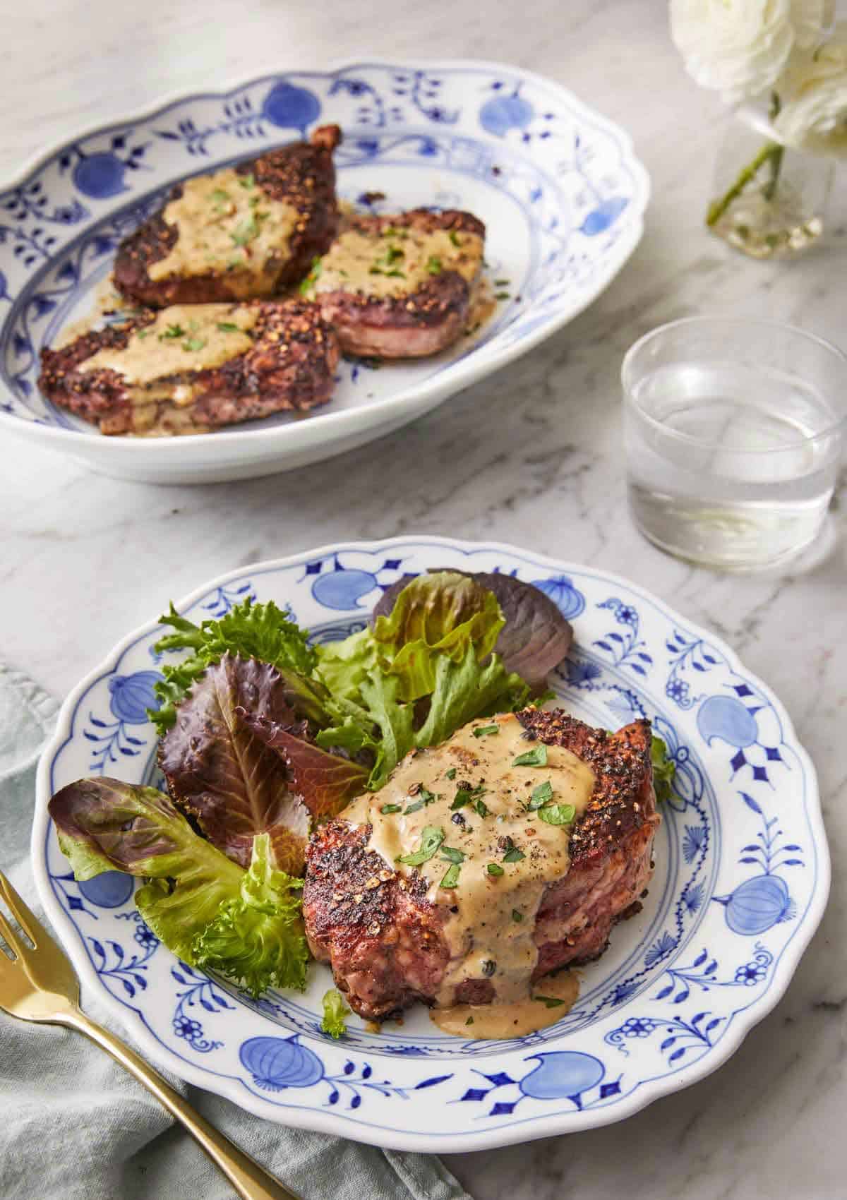 A plate of steak au poivre with a side of salad in front of a platter with three steak au poivre.