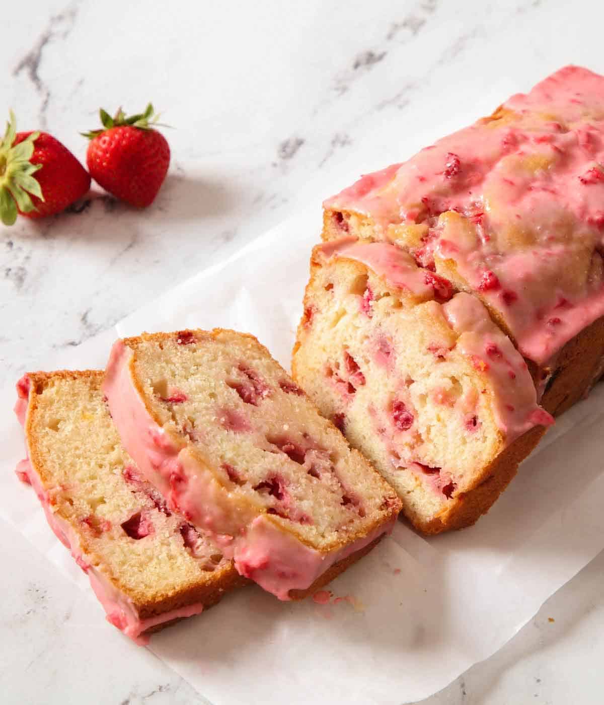 A loaf of strawberry bread with three slices cut in front and two strawberries sitting on the side.