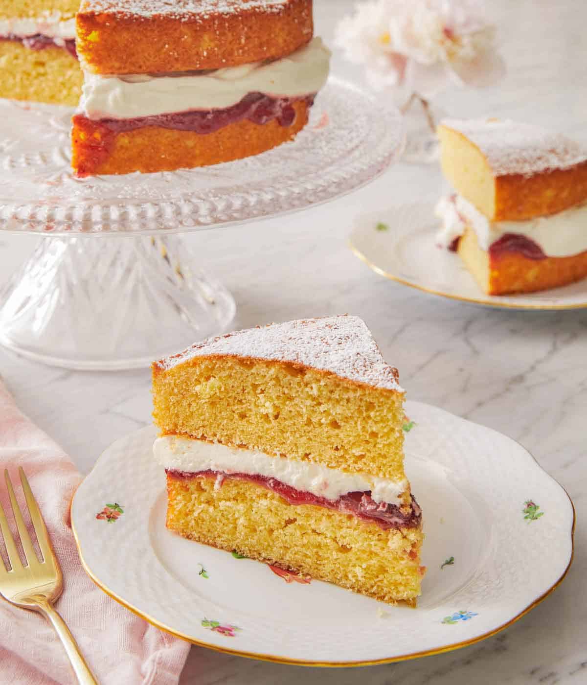 A slice of Victoria sponge cake on a plate in front of a second plate and cake stand.