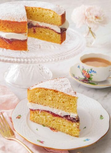 A plate with a slice of Victoria sponge cake in front of a cup of coffee and the rest of the cake.