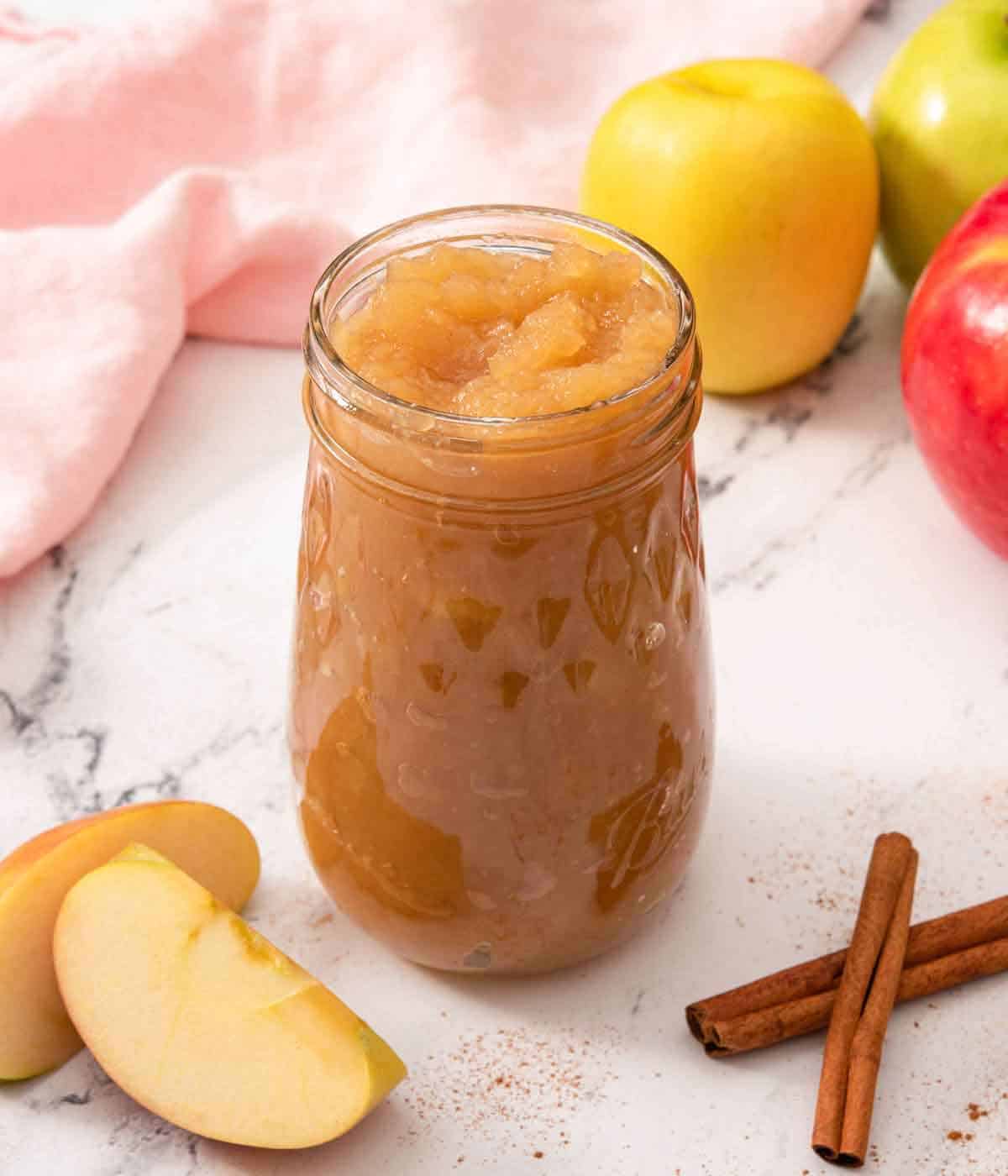 A mason jar filled with applesauce with apples and cinnamon sticks scattered around.