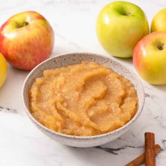 A bowl of apple sauce with multiple apples in the background and some cinnamon sticks scattered around.