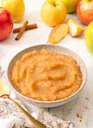 A bowl of apple sauce with fresh apples, some cut, in the background.