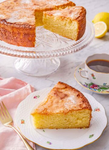 A slice of olive oil cake on a plate in front of the cut cake on a cake stand.