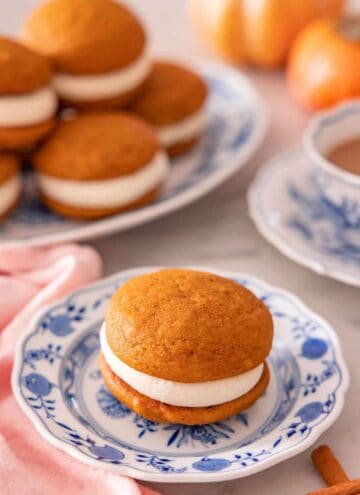 A plate with a pumpkin whoopie pie with additional ones stacked in the back on a platter.