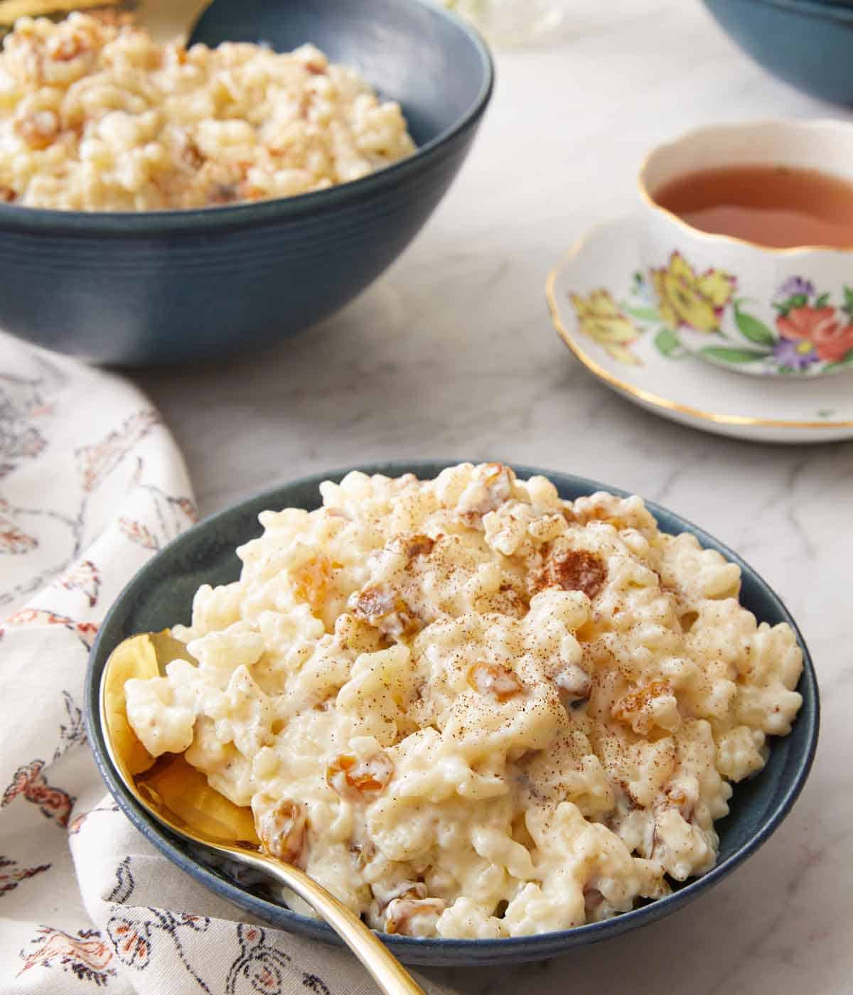 An angled view of a bowl of rice pudding with a golden spoon tucked into the bowl with tea in the background.