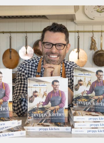 John Kanell in his kitchen behind stacks of the new Preppy Kitchen Cookbook.