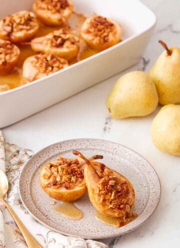 A plate with two baked pears with a baking dish with more in the background and some fresh pears on the side.