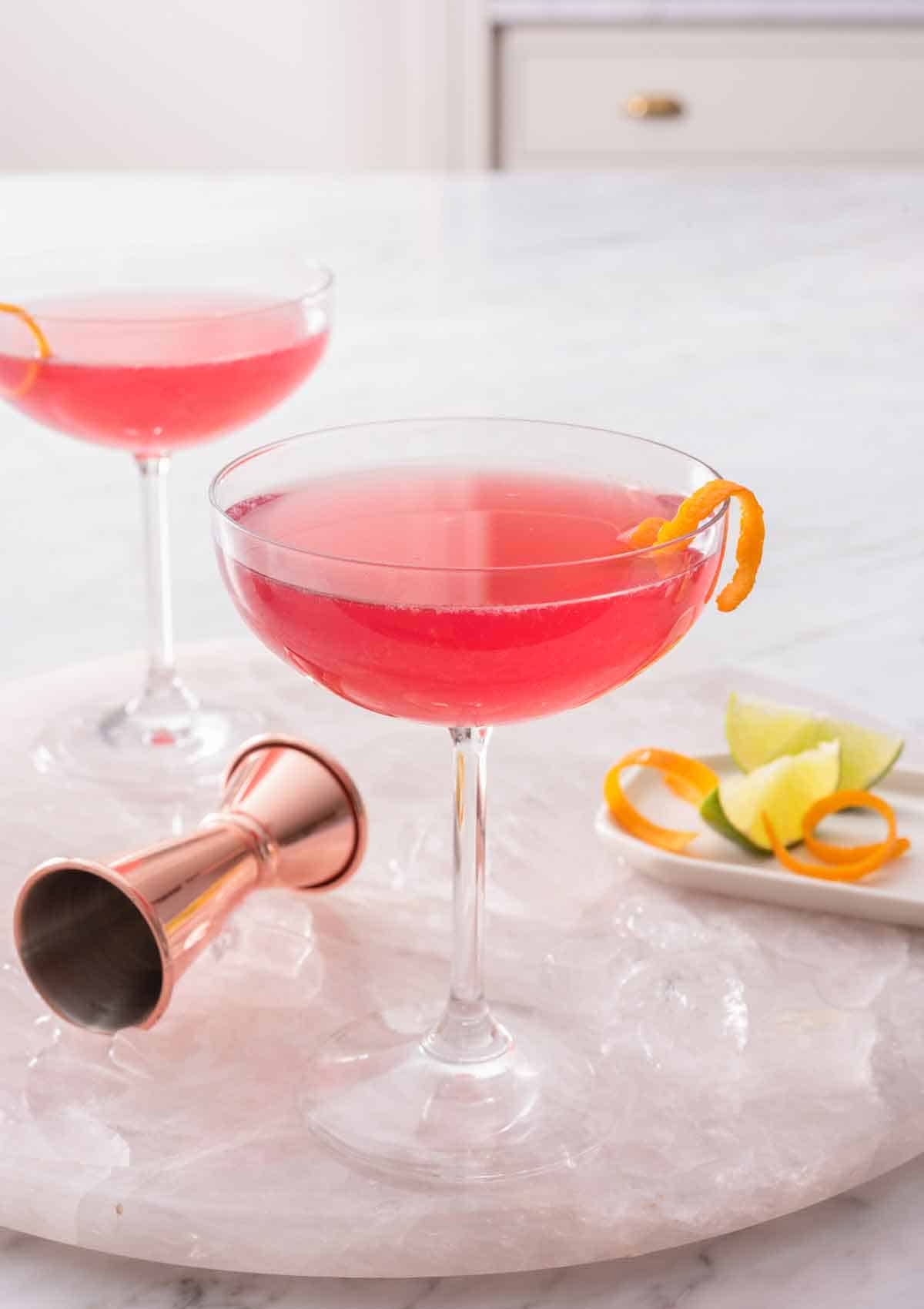Two glasses of Cosmopolitan with an orange garnish on a marble tray with ice and a jigger with more garnishes.