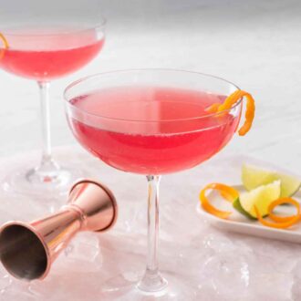 Pinterest graphic of two glasses of Cosmopolitan with one pulled to the front with a jigger, ice, and garnishes scattered around.