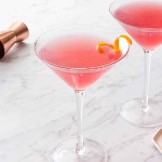Two glasses of Cosmopolitan in martini glasses with garnishes on a plate in front.