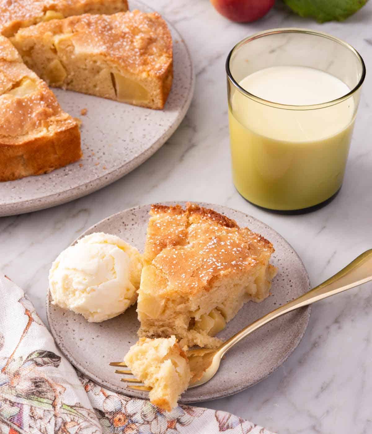 A slice of French apple cake with a forkful in front with a scoop of ice cream beside the cake. A glass of milk and the rest of the cake in the background.