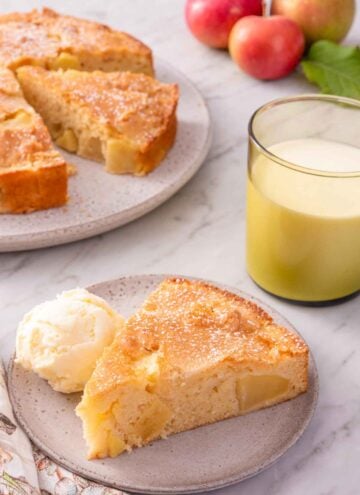 A slice of French apple cake with a scoop of ice cream beside the slice. The rest of the cake, cut, in the background.