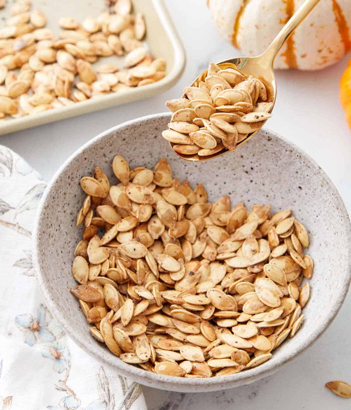 A spoonful of roasted pumpkin seeds scooped up from a bowl of seeds. A sheet pan with more in the background.