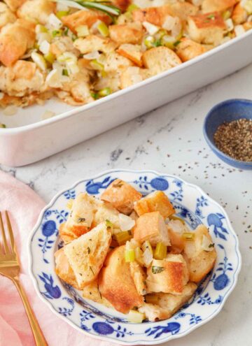 A plate of stuffing with a pinch bowl of pepper and the rest of the baking dish of stuffing in the background.