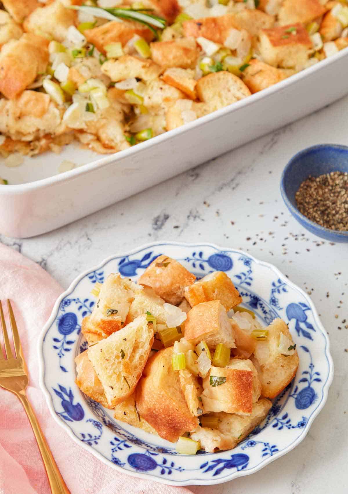 A plate of stuffing with a pinch bowl of pepper and the rest of the baking dish of stuffing in the background.