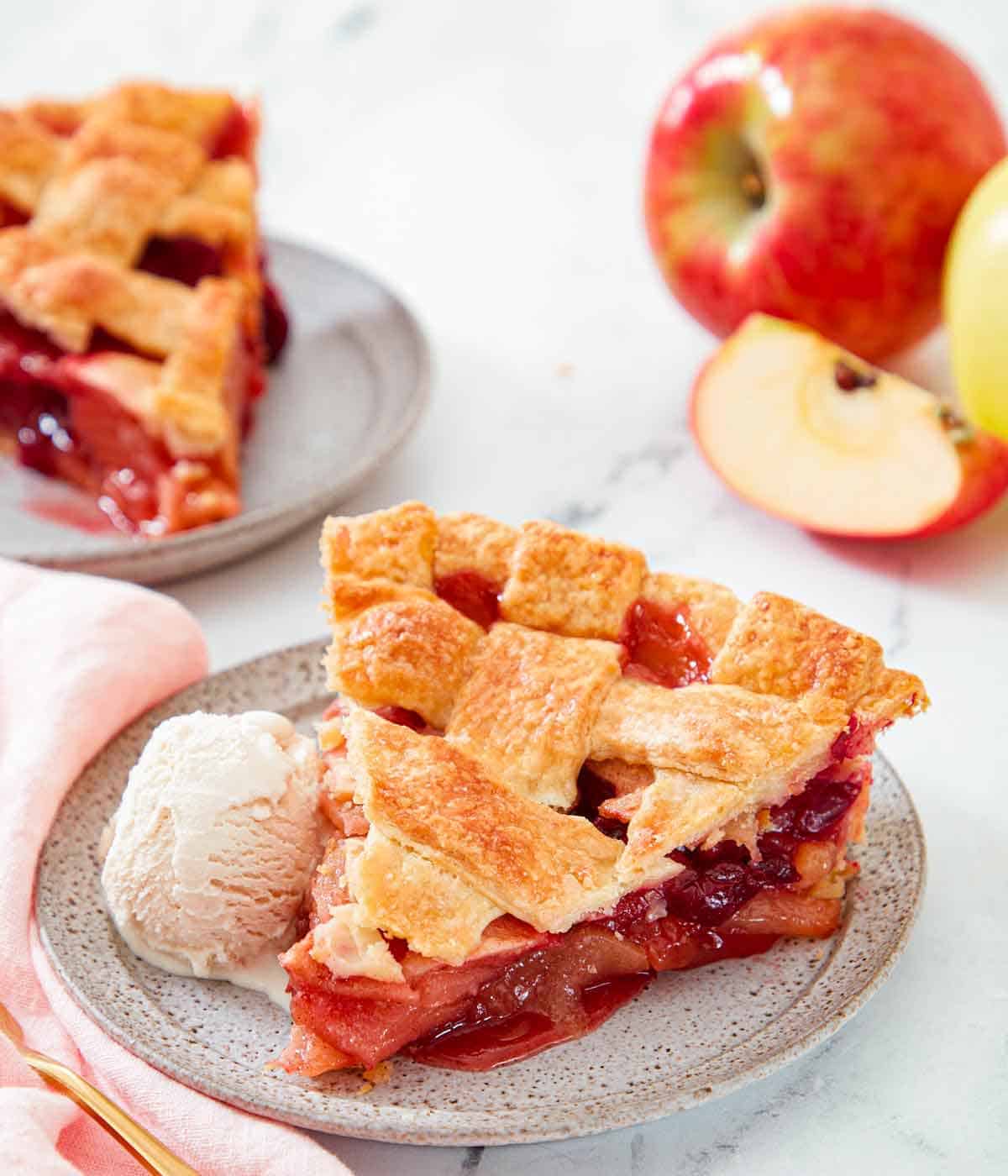 A plate with a slice of apple cranberry pie with a scoop of ice cream. Another plated slice and cut apple in the back.