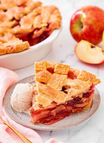 A slice of apple cranberry pie with a scoop of ice cream on the side. Rest of the pie in the background.